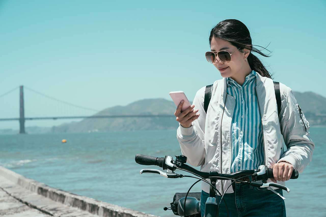 An Asian woman walks her bicycle in front of the Golden Gate Bridge in San Francisco, using her smartphone to search for online information