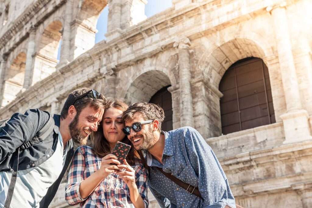Three happy young friends tourists at Colosseum in Rome using smartphone having fun.