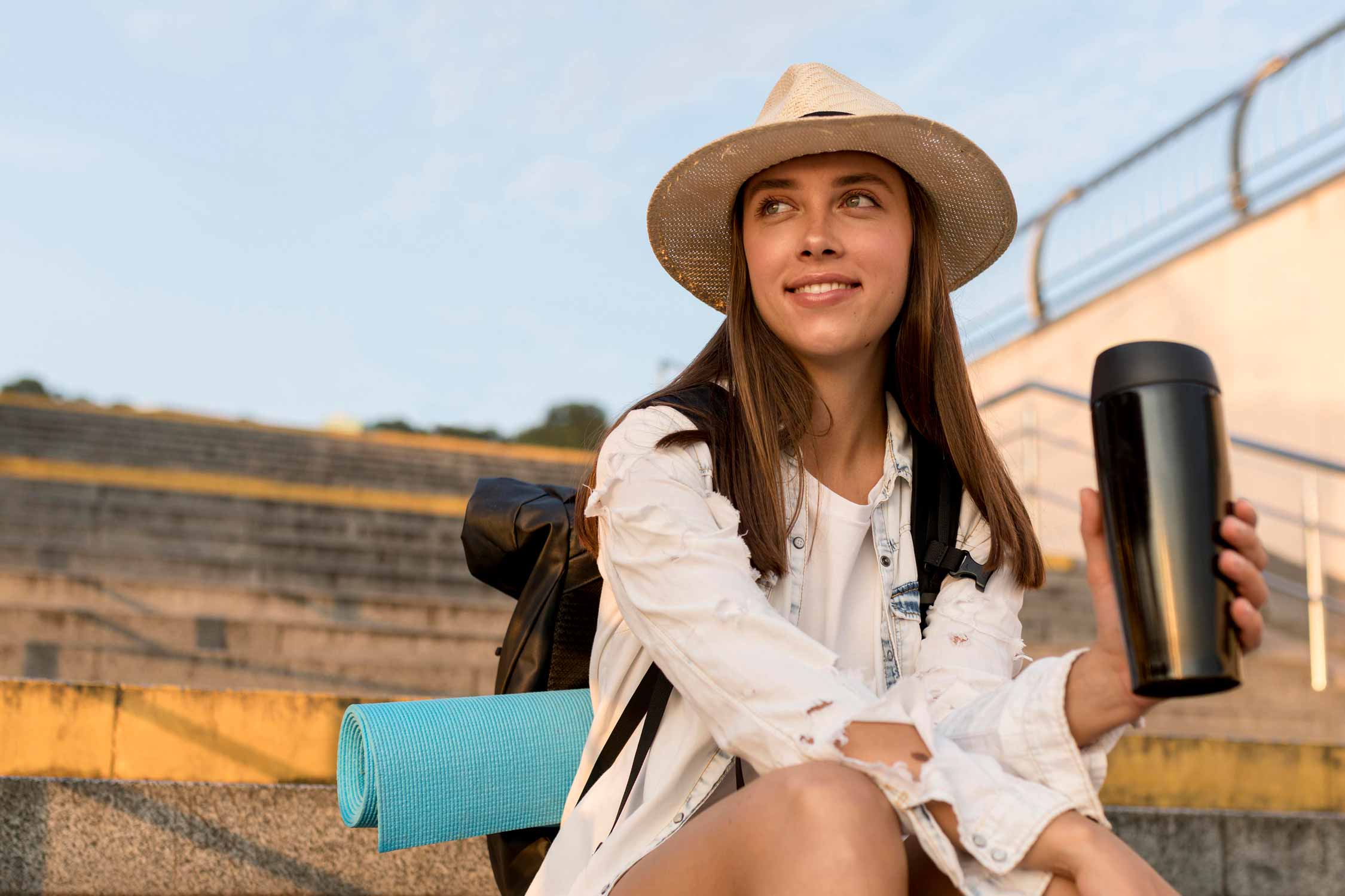 Smiling woman with backpack and hat holding thermos while traveling